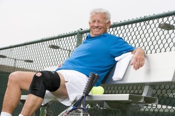 An older man wearing a blue polo shirt and white shorts is sitting on a bench by a tennis court, smiling and relaxing. He has a black knee brace on his right leg, a towel draped over the bench, and two tennis rackets and a tennis ball beside him. A chain-link fence and overhead lights are visible in the background.