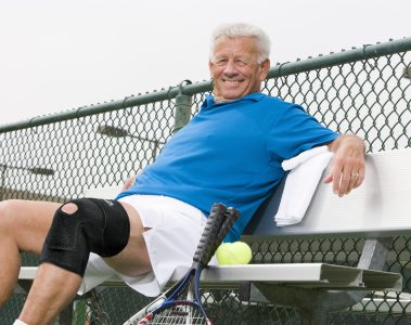 An older man wearing a blue polo shirt and white shorts is sitting on a bench by a tennis court, smiling and relaxing. He has a black knee brace on his right leg, a towel draped over the bench, and two tennis rackets and a tennis ball beside him. A chain-link fence and overhead lights are visible in the background.